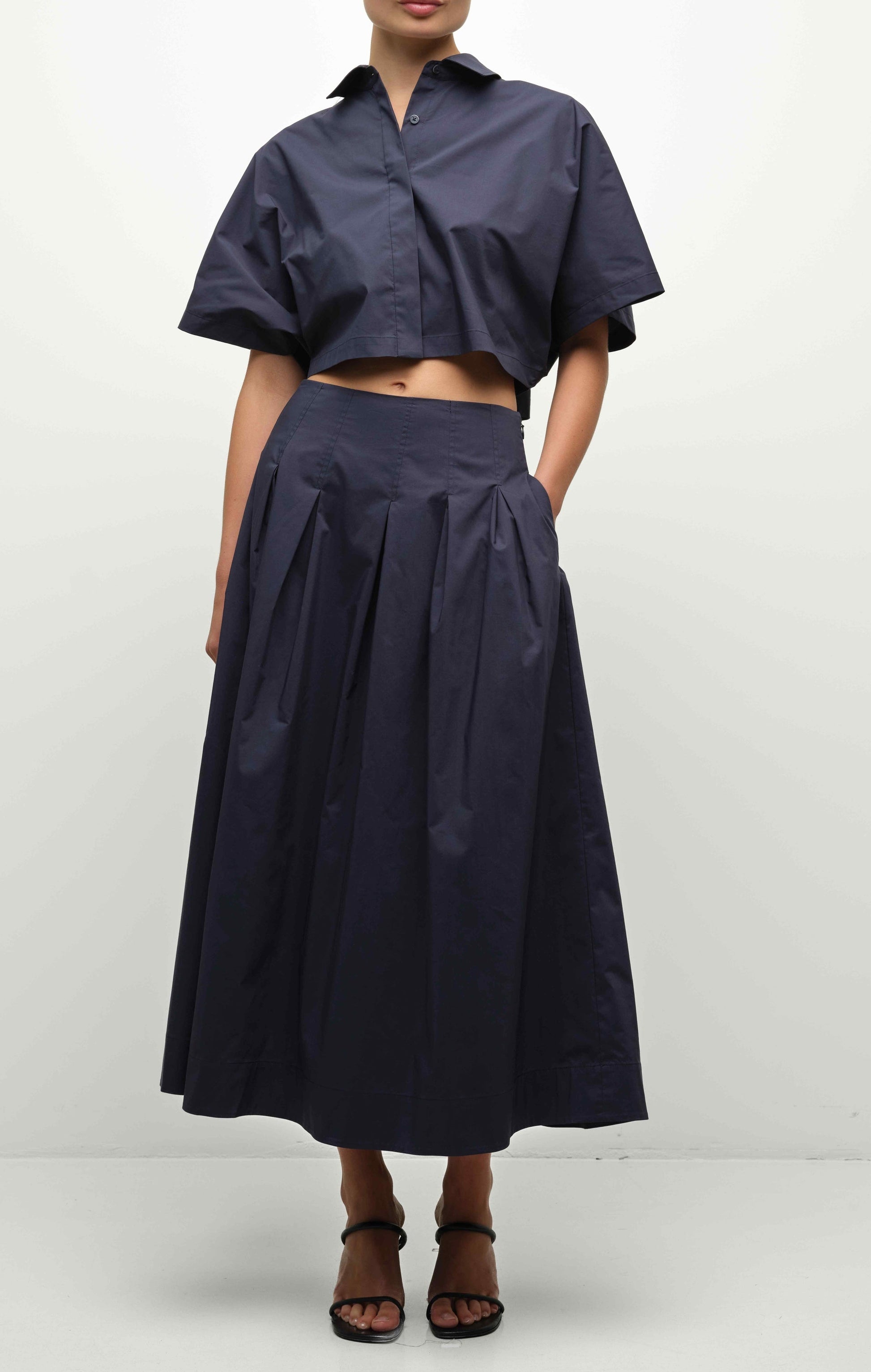 A person wears the Simkhai Elora Midi Skirt with a matching dark navy short-sleeve cropped button-up shirt and black sandals, standing against a plain background.