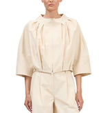 A person wearing the Simkhai Jess Pleated Jacket, a loose-fitting, cream-colored zip-up blouse with three-quarter sleeves, paired with matching high-waisted pants.