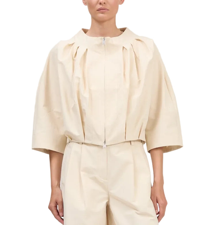 A person wearing the Simkhai Jess Pleated Jacket, a loose-fitting, cream-colored zip-up blouse with three-quarter sleeves, paired with matching high-waisted pants.
