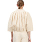 A person is seen from the back wearing the Simkhai Jess Pleated Jacket in cream, a loose-fitting, pleated top with three-quarter sleeves, paired with matching pants. Hair is styled in a low, neat ponytail.
