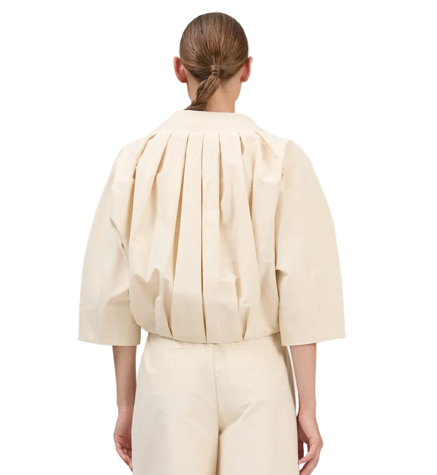 A person is seen from the back wearing the Simkhai Jess Pleated Jacket in cream, a loose-fitting, pleated top with three-quarter sleeves, paired with matching pants. Hair is styled in a low, neat ponytail.