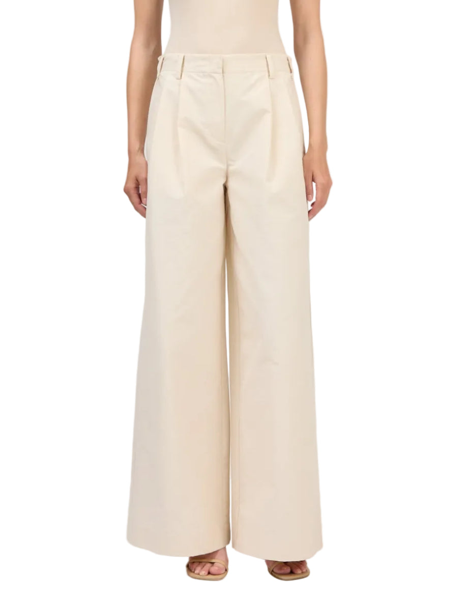 A person wears the Simkhai Leroy Pleated Wide Leg Pant—high-waisted beige trousers with front pleats and belt loops—paired with open-toe sandals.