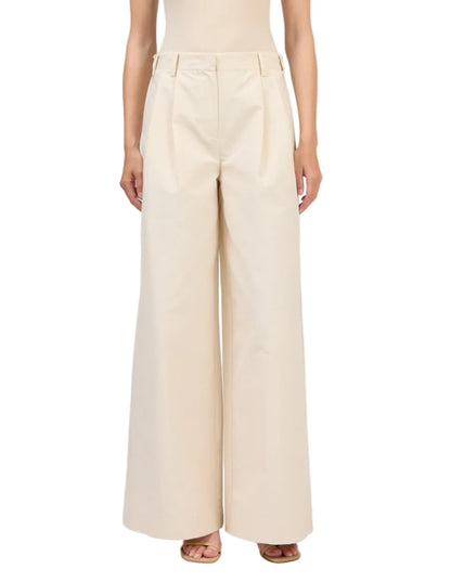 A person wears the Simkhai Leroy Pleated Wide Leg Pant—high-waisted beige trousers with front pleats and belt loops—paired with open-toe sandals.