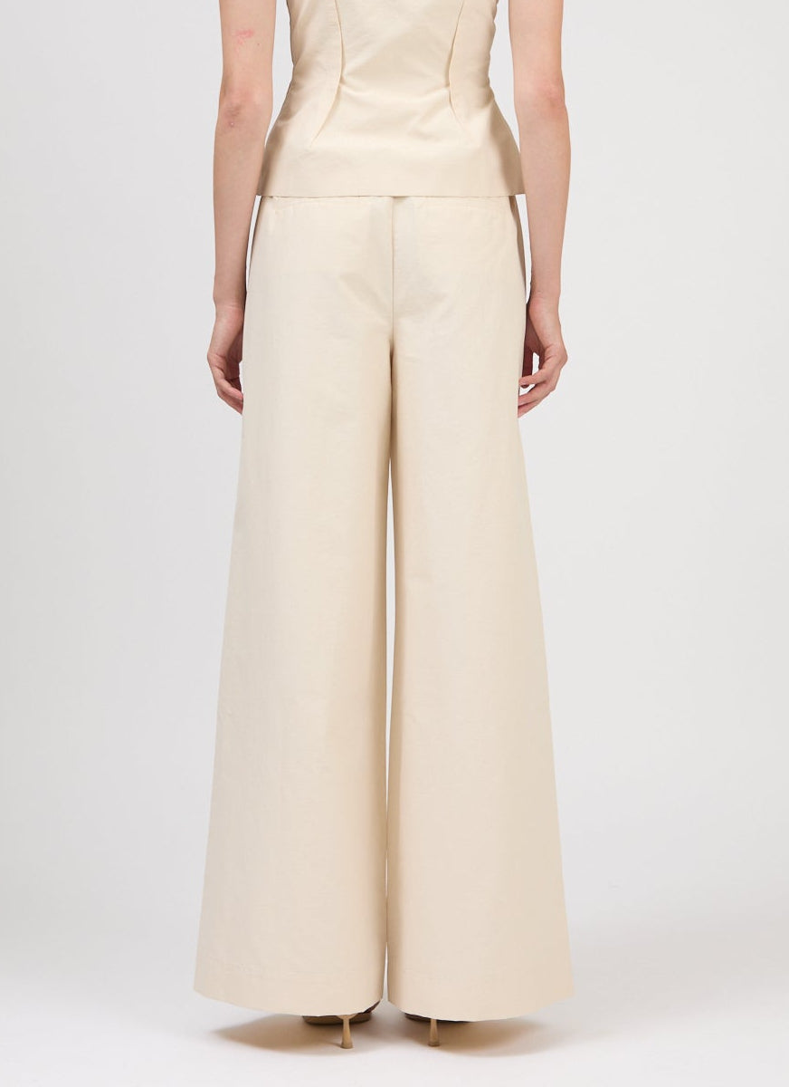 Back view of a person wearing Simkhai's Leroy Pleated Wide Leg Pant in cream, paired with a matching fitted top, standing against a plain white background.
