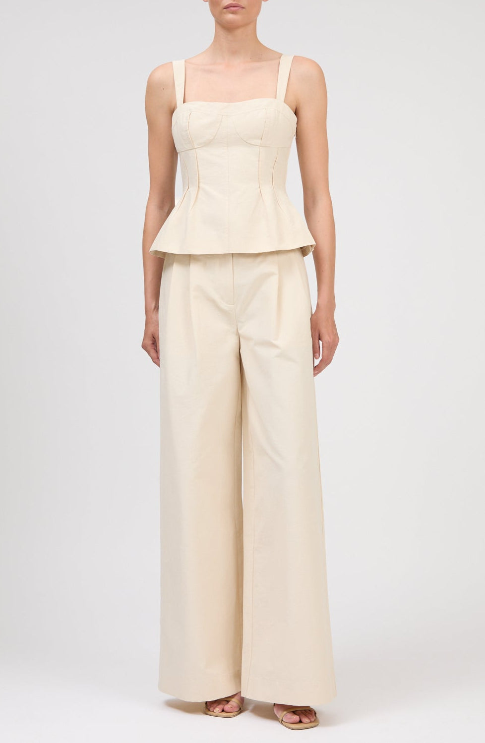 A person wearing Simkhai's Leroy Pleated Wide Leg Pant paired with a sleeveless, cream peplum top and open-toe sandals, standing against a plain background.