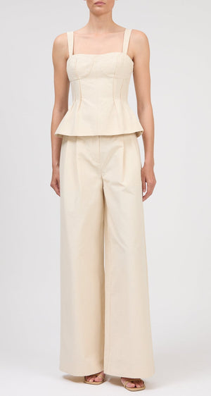 A person wears the Simkhai April Bustier Top, a sleeveless fitted beige peplum top, paired with matching wide-leg pants and open-toe heels, standing against a plain background.