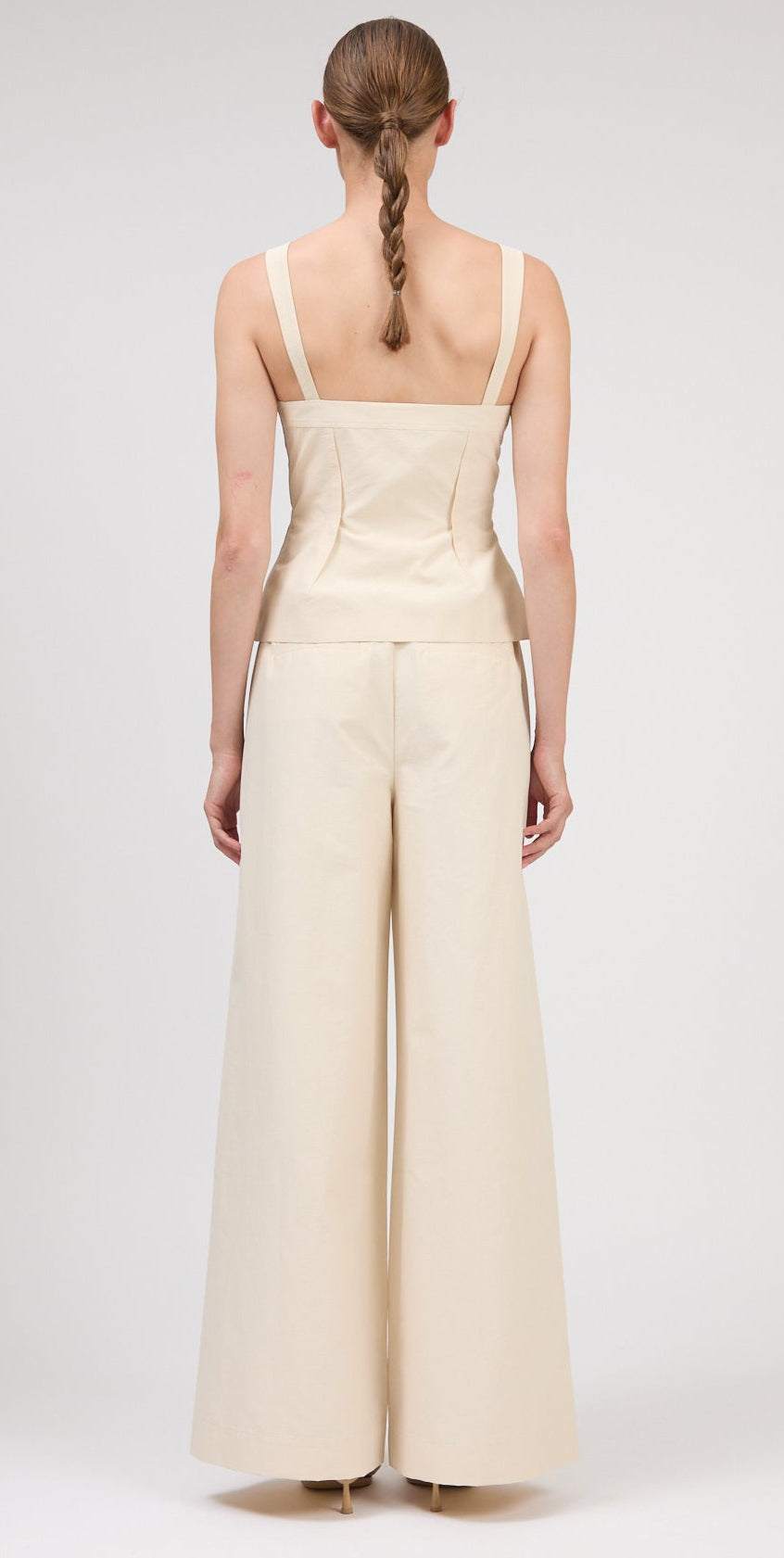 A person with a single braid is shown from the back, wearing the Simkhai April Bustier Top in beige with wide straps, paired with matching high-waisted, wide-leg pants.