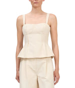 A woman wearing the Simkhai April Bustier Top, a cream sleeveless peplum style with wide straps, paired with matching high-waisted pants, stands against a white background.