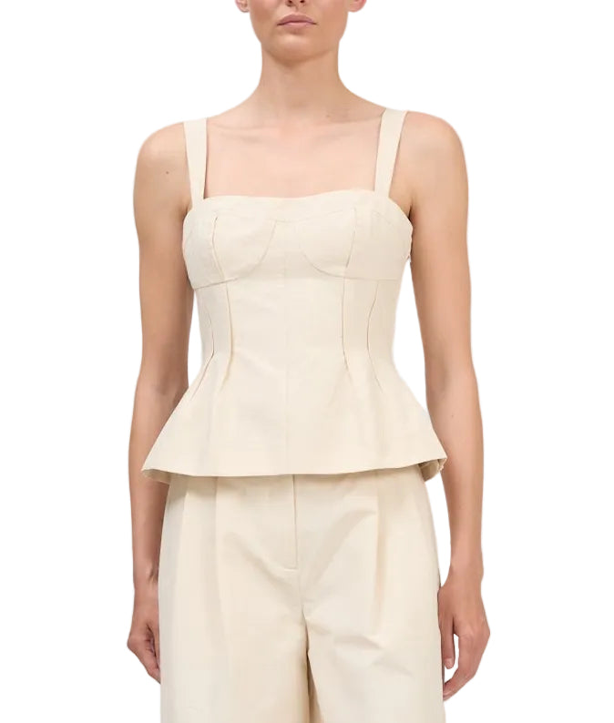 A woman wearing the Simkhai April Bustier Top, a cream sleeveless peplum style with wide straps, paired with matching high-waisted pants, stands against a white background.
