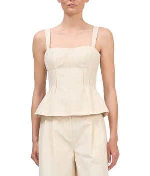 A woman wearing the Simkhai April Bustier Top, a cream sleeveless peplum style with wide straps, paired with matching high-waisted pants, stands against a white background.