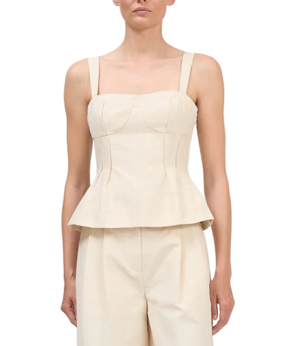 A woman wearing the Simkhai April Bustier Top, a cream sleeveless peplum style with wide straps, paired with matching high-waisted pants, stands against a white background.