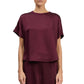 A person wearing the Simkhai Addy Satin T-Shirt—a loose-fitting, short-sleeve burgundy top with a round neckline—paired with matching pants, standing against a white background.