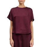 A person wearing the Simkhai Addy Satin T-Shirt—a loose-fitting, short-sleeve burgundy top with a round neckline—paired with matching pants, standing against a white background.