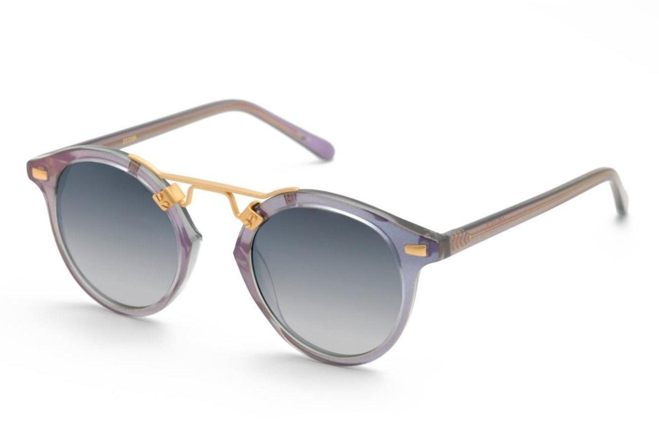 The Krewe St Louis Mirrored Sunglasses feature round luxury acetate frames with purple accents, gray-tinted gradient lenses, and 24K gold-plated detailing on the temples and bridge, offering complete UVA/UVB protection.