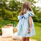 A young girl stands on a picnic blanket outdoors, surrounded by pillows and greenery, wearing The Beaufort Bonnet Company Baby Girls’ and Girls’ Mary Dal Dress in Audubon Park Plaid with a large bow, facing away.
