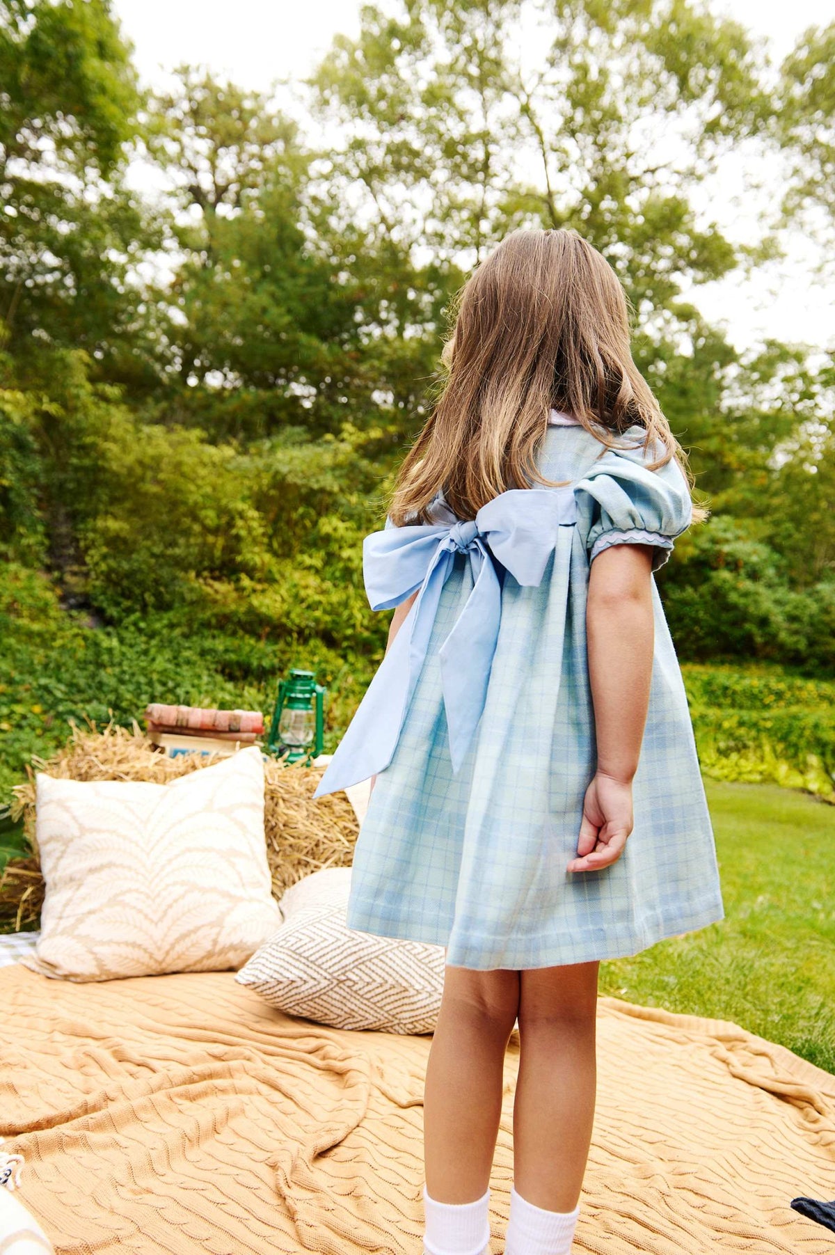 A young girl stands on a picnic blanket outdoors, surrounded by pillows and greenery, wearing The Beaufort Bonnet Company Baby Girls’ and Girls’ Mary Dal Dress in Audubon Park Plaid with a large bow, facing away.