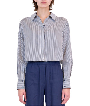 A person models the Simkhai Renata Stripe Cropped Button Down Shirt in blue and white stripes with long sleeves, paired with navy pants, standing against a plain white background.