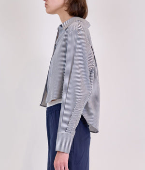 A person wears the Simkhai Renata Stripe Cropped Button Down Shirt—blue and white vertical stripes with long sleeves—paired with navy pants, shown from the side against a plain background to highlight its tailored, versatile fit.