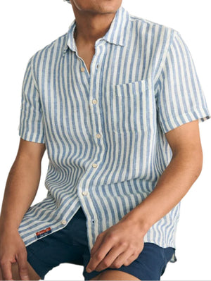 A person is seated, wearing Faherty's bestseller, the Short-Sleeve Palma Linen Shirt with blue and white stripes, paired with dark shorts. One hand rests casually on their knee.