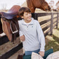 A boy in a light blue Boys’ Prepletic Hayword Half Zip by The Beaufort Bonnet Company stands by a wooden fence, with a horse, saddle, chair, and patterned pillow nearby.