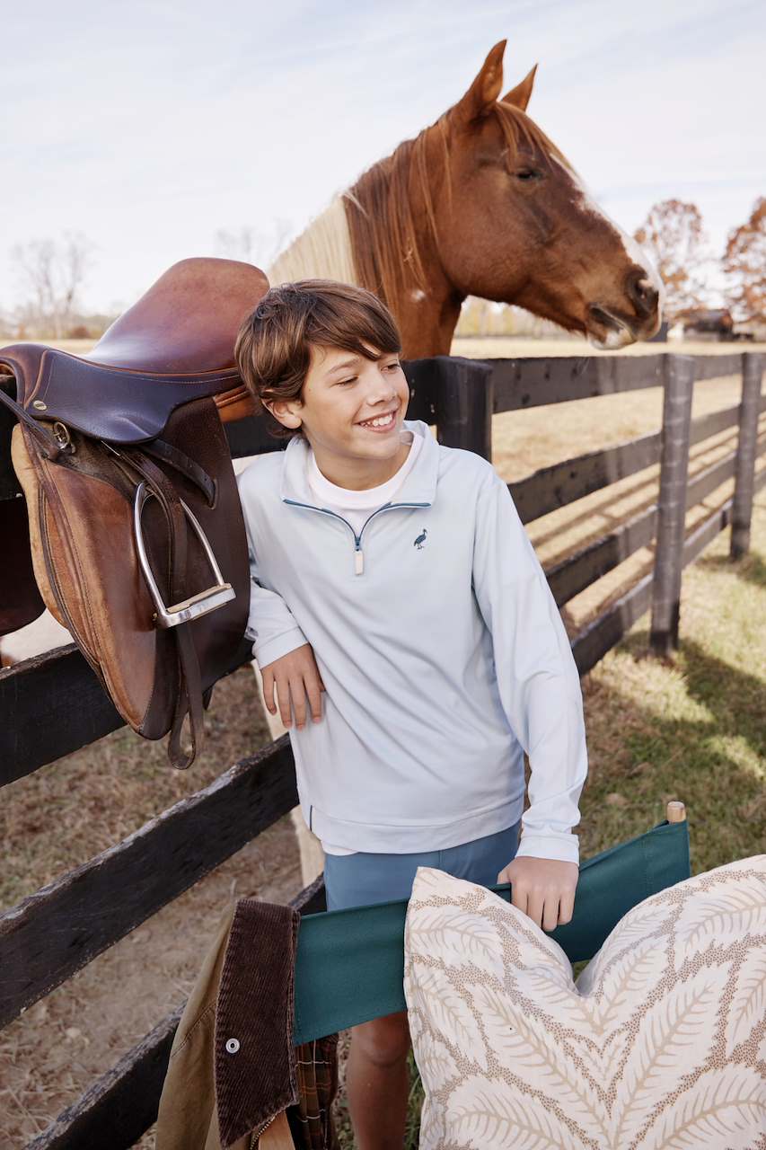 A boy in a light blue Boys’ Prepletic Hayword Half Zip by The Beaufort Bonnet Company stands by a wooden fence, with a horse, saddle, chair, and patterned pillow nearby.