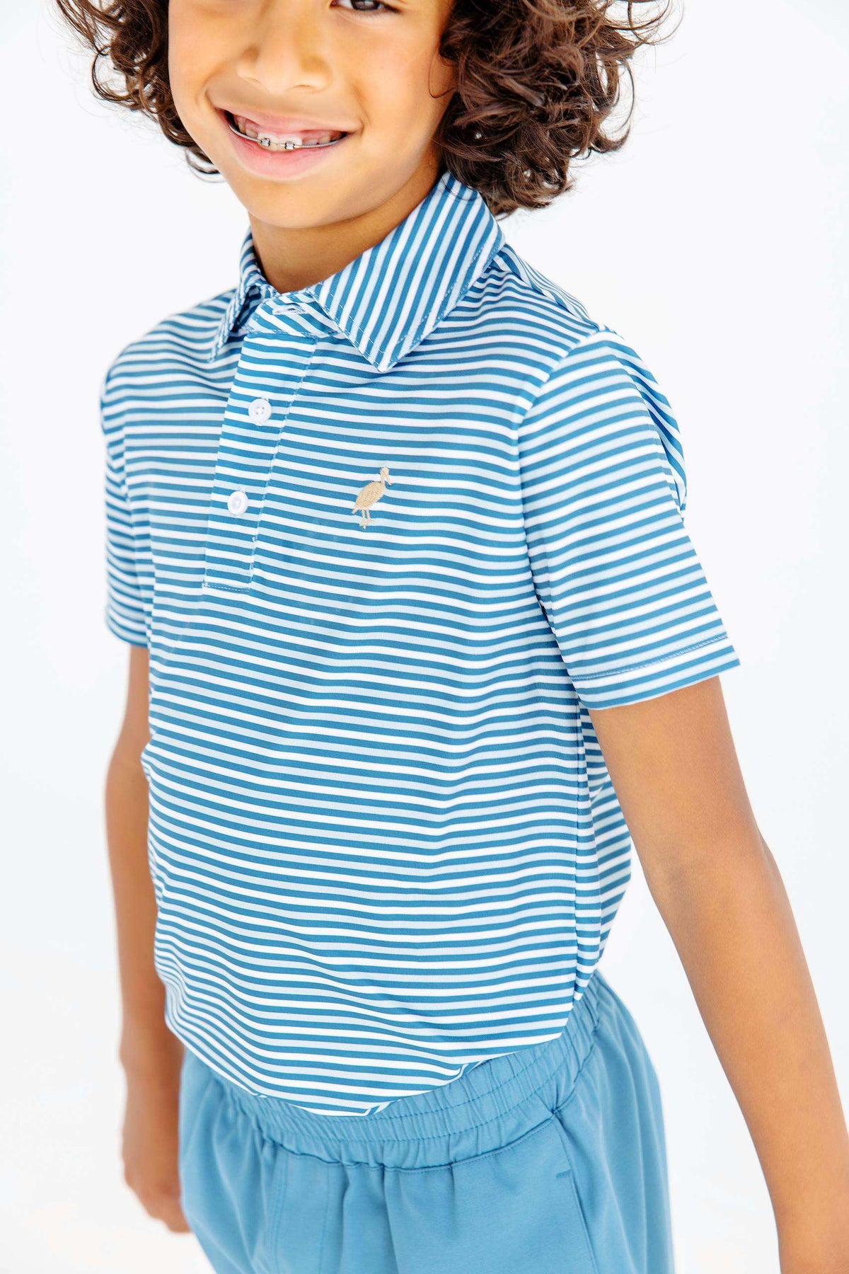 Smiling child wearing The Beaufort Bonnet Company Boys’ Prepletic™ Prim & Proper Polo, showcasing blue and white stripes with short sleeves, paired with light blue pants, standing against a plain white background.