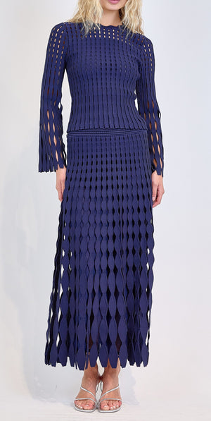 A woman models the Simkhai Wilma Skirt, a navy blue pointelle knit design with an openwork diamond pattern, paired with silver sandals against a plain background.