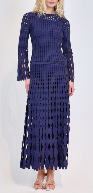 A person wears the Simkhai Peyton Top, a long-sleeved, navy blue pointelle knit top with diamond-shaped cutouts, paired with silver sandals against a plain background.