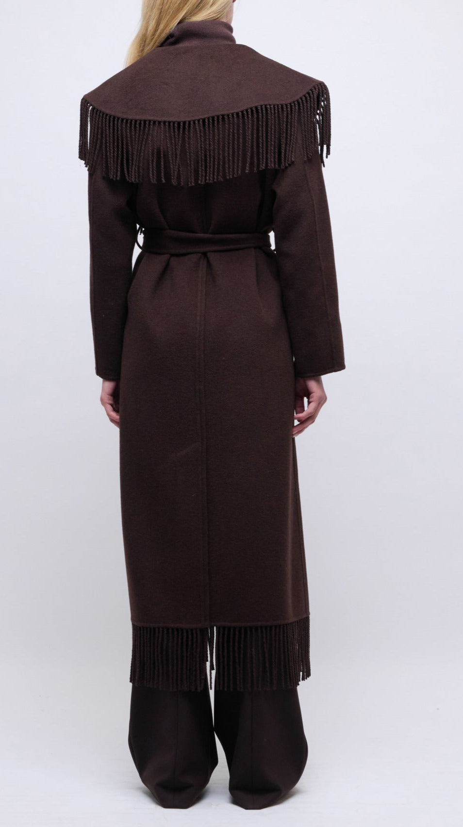 A person is seen from the back wearing the Simkhai Carrie Fringe Robe Coat, a long brown coat with a fringed hem and shoulder fringe details, set against a plain white background.