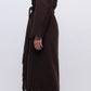 A person stands sideways against a plain background wearing the Simkhai Carrie Fringe Robe Coat in dark brown, featuring a self-tie belt and fringed hem, paired with dark pants and black pointed shoes.