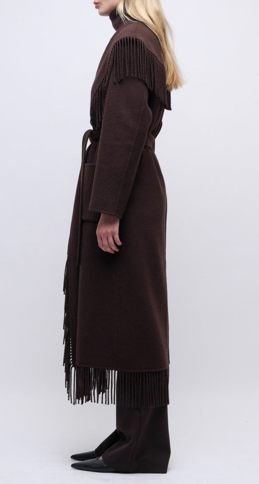 A person stands sideways against a plain background wearing the Simkhai Carrie Fringe Robe Coat in dark brown, featuring a self-tie belt and fringed hem, paired with dark pants and black pointed shoes.