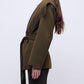 A woman in side profile wears the Simkhai Rowen Fringe Jacket in olive green, featuring a belted waist and fringe-detailed shoulders, paired with dark pants for a subtle Western-inspired look against a plain light background.