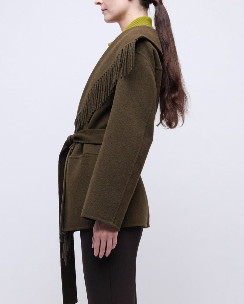 A woman in side profile wears the Simkhai Rowen Fringe Jacket in olive green, featuring a belted waist and fringe-detailed shoulders, paired with dark pants for a subtle Western-inspired look against a plain light background.