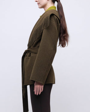 A woman in side profile wears the Simkhai Rowen Fringe Jacket in olive green, featuring a belted waist and fringe-detailed shoulders, paired with dark pants for a subtle Western-inspired look against a plain light background.