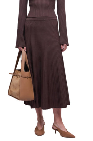 A person wearing the Simkhai Chiara Midi Skirt with a matching long-sleeved, ribbed brown top, holding a tan handbag and wearing tan heeled mules.