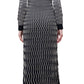 A woman stands with her back to the camera, wearing the Simkhai Kassidy Polo Midi Dress—an ankle-length black dress with a white wavy line pattern—and black sandals.