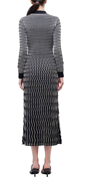 A woman stands with her back to the camera, wearing the Simkhai Kassidy Polo Midi Dress—an ankle-length black dress with a white wavy line pattern—and black sandals.