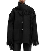 A person wears Toteme's Fringed Scarf Jacket—a black wool-blend bouclé coat with oversized fit, large buttons, and fringe details on the sleeves, hem, and scarf—paired with a small black clutch.