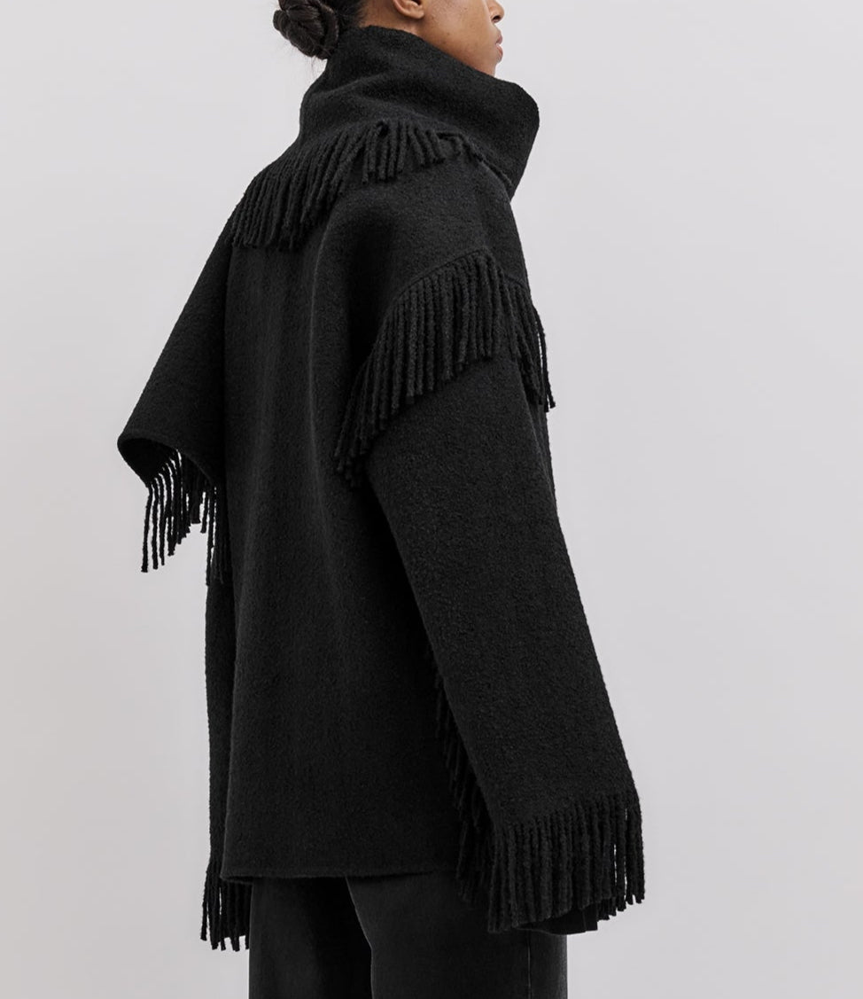 Person wearing Toteme's Fringed Scarf Jacket, a black wool-blend bouclé coat with fringe on the sleeves, back, and hem, featuring an oversized silhouette against a plain light background.