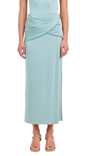 A person wearing the Simkhai Vienna Midi Skirt in light blue, featuring a figure-flattering fit and crisscross waist detail, paired with beige sandals; shown from the shoulders down.