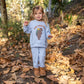 A young child stands outdoors on fallen leaves, surrounded by trees and foliage, wearing Feather 4 Arrow’s Baby and Kids’ Hudson Hacci Jogger in light blue with a playful bear graphic sweatshirt.