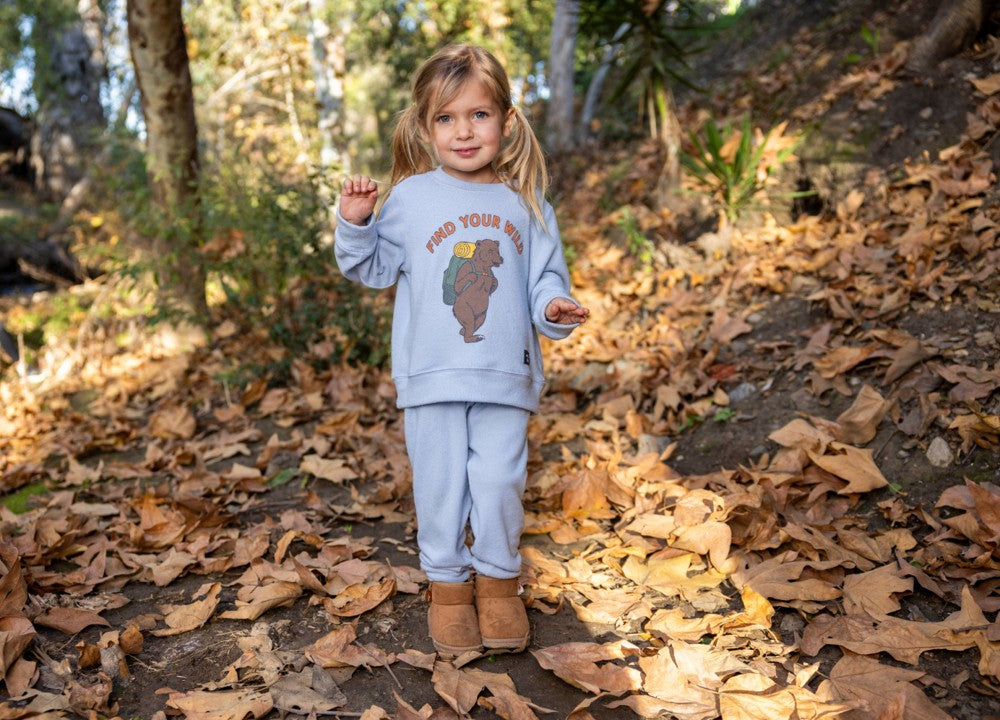 A young child stands outdoors on fallen leaves, surrounded by trees and foliage, wearing Feather 4 Arrow’s Baby and Kids’ Hudson Hacci Jogger in light blue with a playful bear graphic sweatshirt.