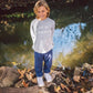 A child with blond hair stands on a tree stump near a stream, wearing a gray “LOCAL COWBOY” sweatshirt, Feather 4 Arrow’s Baby and Kids’ Hudson Hacci Jogger in blue, and white shoes. Fallen leaves cover the ground and the water’s surface.