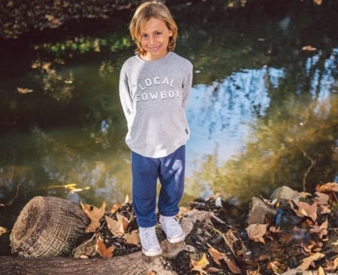 A child with blond hair stands on a tree stump near a stream, wearing a gray “LOCAL COWBOY” sweatshirt, Feather 4 Arrow’s Baby and Kids’ Hudson Hacci Jogger in blue, and white shoes. Fallen leaves cover the ground and the water’s surface.