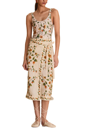 A woman models the Luna Midi Pareo by Agua By Aguabendita, standing on draped cloth in a gymnasium, framed by a stage and gold curtains in the background.