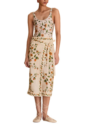 A woman wearing the Gema One Piece by Agua By Aguabendita, made from recycled polyester, stands on a cloth-covered platform in an empty gymnasium with a stage and gold curtains behind her.