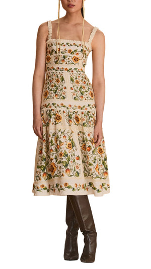 A woman wearing the Nispero Dress by Agua By Aguabendita, a pure linen floral midi with a square neckline, stands on a draped platform in the center of an empty gym, framed by wooden floors and a gold curtain backdrop.