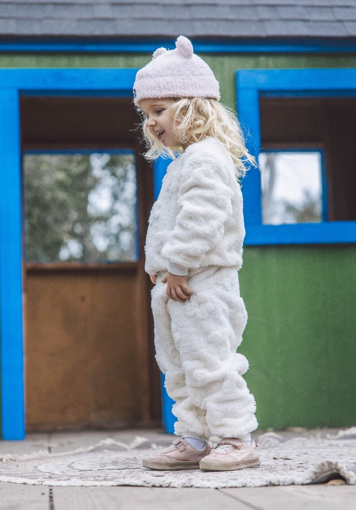 A young child in a Feather 4 Arrow Baby Girls’ and Girl’s Yummy Plush Set and a pink hat stands outside a colorful playhouse with green walls and blue trim.