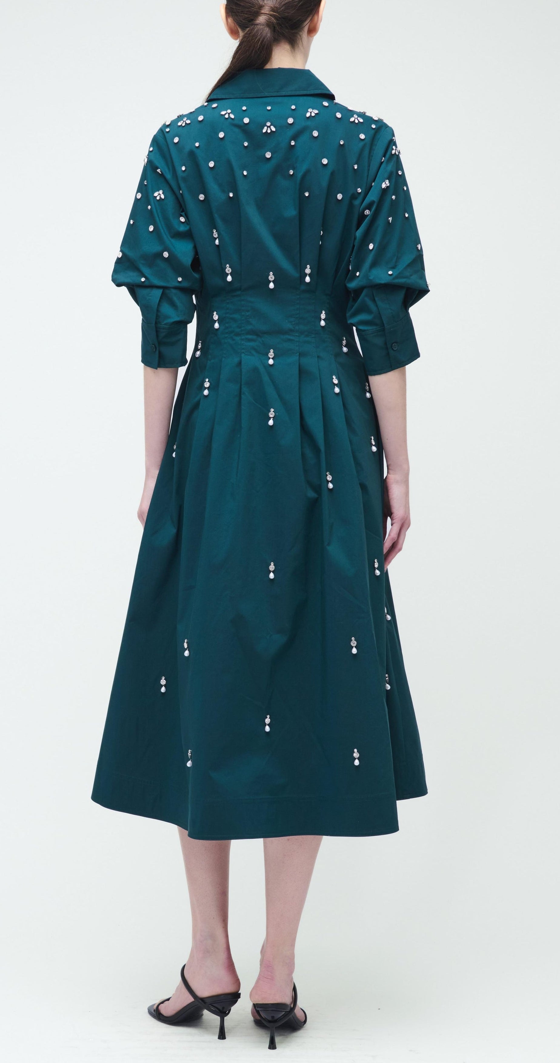 A woman faces away, wearing the Simkhai Jazz Embellished Midi Dress in teal cotton poplin with white embroidered dots, an A-line skirt, three-quarter sleeves, and black heeled sandals.