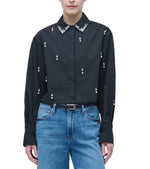 A person wearing the Simkhai Renata Embellished Cropped Shirt with small embroidered details and a beaded collar, paired with blue jeans and a black belt.